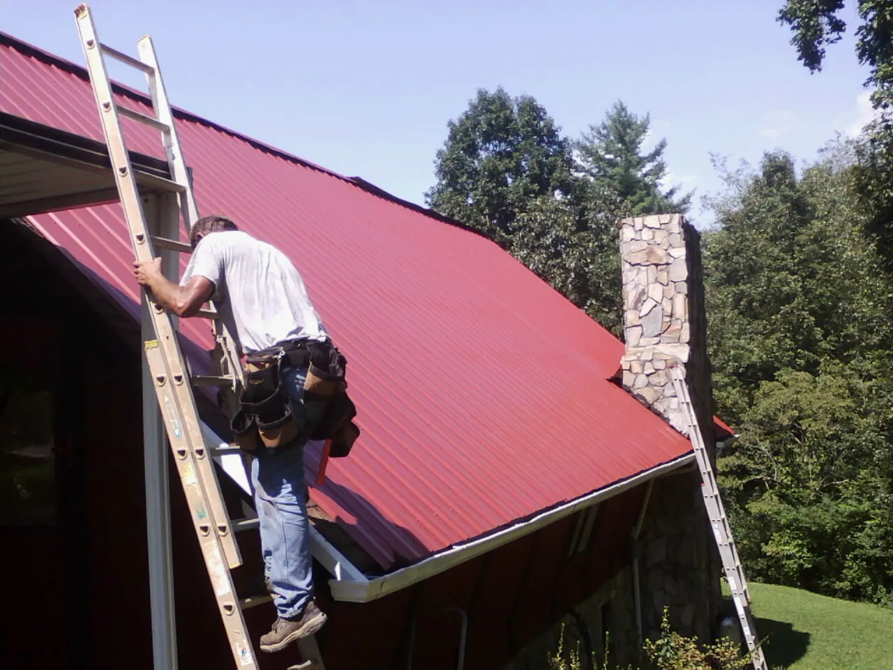 Roofer on a ladder beside a red metal roof and stone chimney — Soffit & Fascia Repair by Roofing Brothers in Spring City